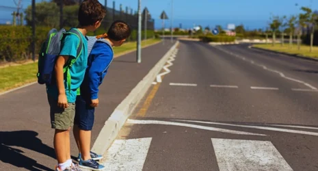 Two schoolboys standing at the edge of a crosswalk, looking to their left as if checking for traffic before crossing the road.