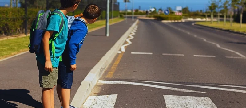 Two schoolboys standing at the edge of a crosswalk, looking to their left as if checking for traffic before crossing the road.
