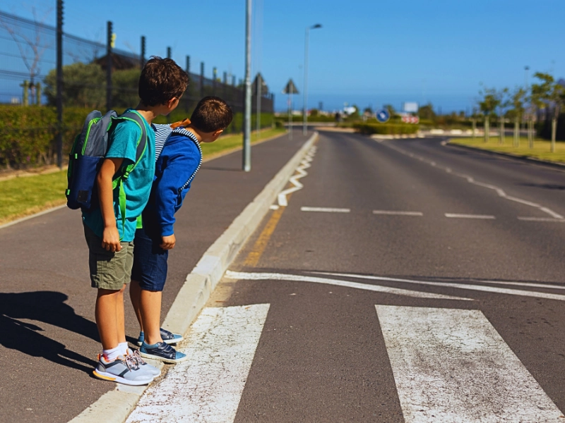 Two schoolboys standing at the edge of a crosswalk, looking to their left as if checking for traffic before crossing the road.