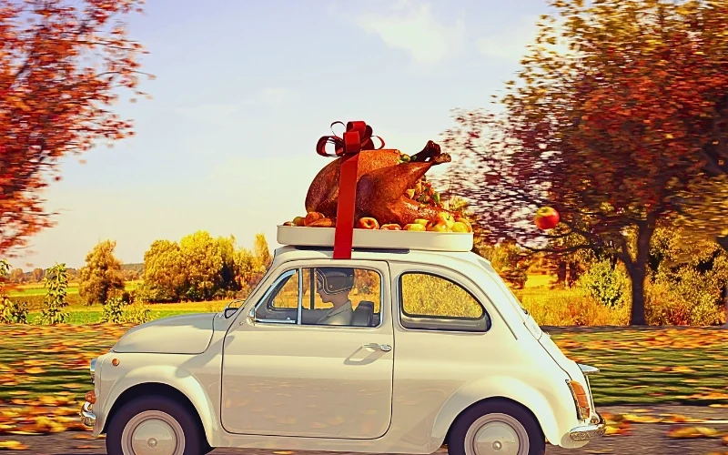a small vintage car driving through an autumn landscape with colorful fall trees. On the car's roof is a large roasted turkey decorated with a red ribbon and bow, surrounded by small fruits or vegetables. The scene suggests a festive, holiday atmosphere with warm lighting and fallen leaves.