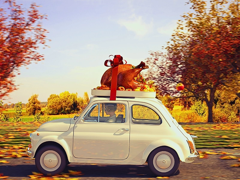 a small vintage car driving through an autumn landscape with colorful fall trees. On the car's roof is a large roasted turkey decorated with a red ribbon and bow, surrounded by small fruits or vegetables. The scene suggests a festive, holiday atmosphere with warm lighting and fallen leaves.