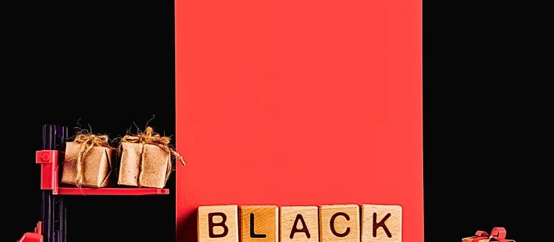 a red and black forklift carrying two small, wrapped gift boxes against a black and red background. In front of the forklift, wooden blocks spell out "BLACK FRIDAY," and a single wrapped gift box sits to the right.