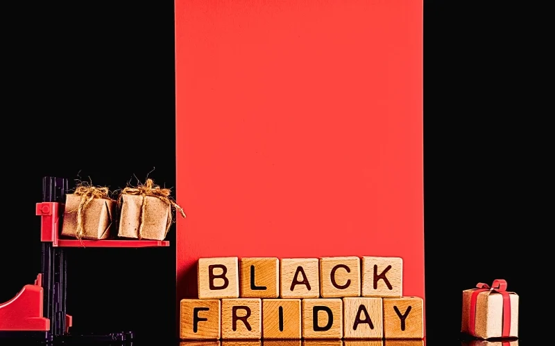 a red and black forklift carrying two small, wrapped gift boxes against a black and red background. In front of the forklift, wooden blocks spell out "BLACK FRIDAY," and a single wrapped gift box sits to the right.