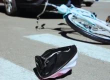 A pink child's helmet lies in the street at the pedestrian crossing; in the background, a bicycle is seen on the ground along with a car that appears to have hit the bicycle. Bicycle Accidents Often Lead to Serious Injuries