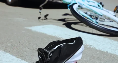 A pink child's helmet lies in the street at the pedestrian crossing; in the background, a bicycle is seen on the ground along with a car that appears to have hit the bicycle. Bicycle Accidents Often Lead to Serious Injuries