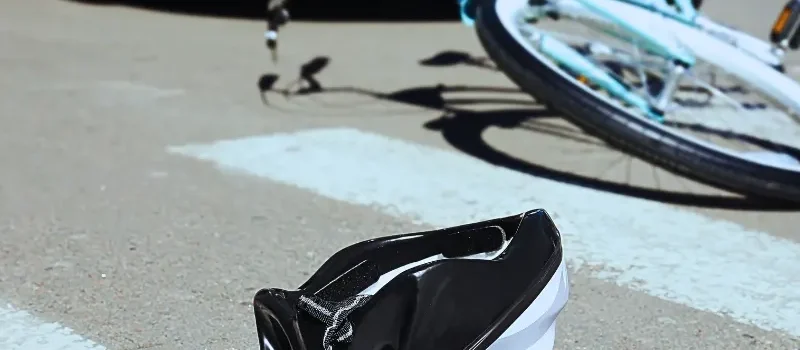 A pink child's helmet lies in the street at the pedestrian crossing; in the background, a bicycle is seen on the ground along with a car that appears to have hit the bicycle. Bicycle Accidents Often Lead to Serious Injuries