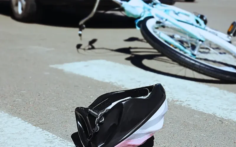 A pink child's helmet lies in the street at the pedestrian crossing; in the background, a bicycle is seen on the ground along with a car that appears to have hit the bicycle. Bicycle Accidents Often Lead to Serious Injuries