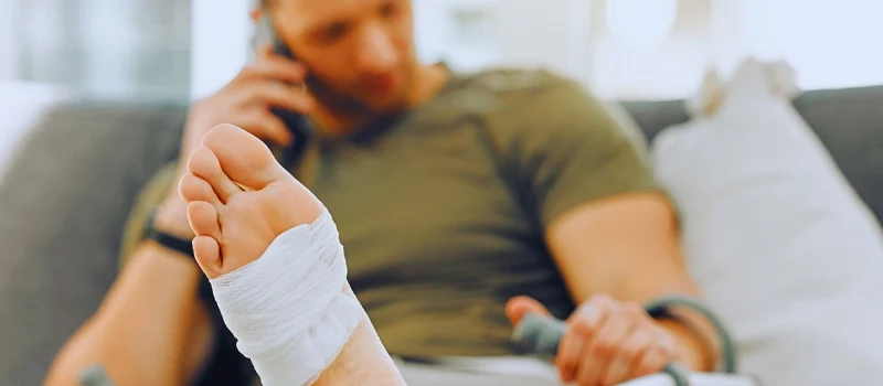 Man with bandaged foot talking on phone while holding crutch. file a personal injury claim after leaving Florida