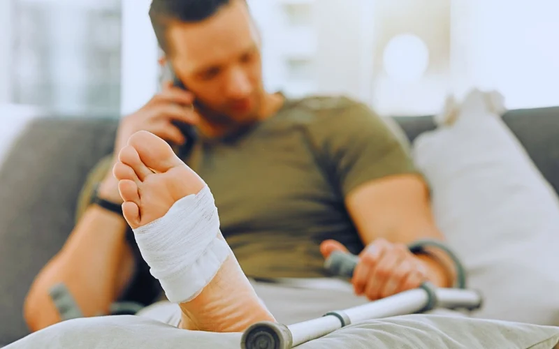 Man with bandaged foot talking on phone while holding crutch. file a personal injury claim after leaving Florida