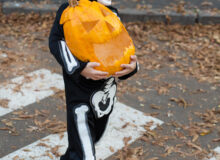 a child dressed as a skeleton carrying a pumpkin while crossing the street